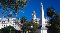 Blick auf die belebte Plaza de Mayo mit umliegenden Häusern und einem Denkmal in der Mitte des Platzes.