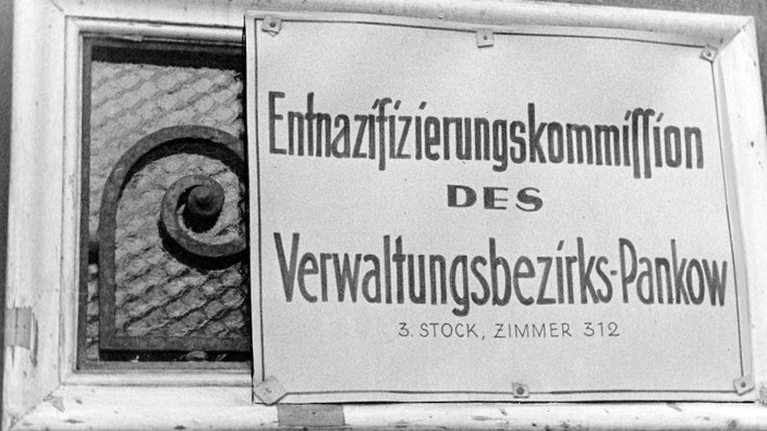 Auf einem Schild steht „Entnazifizierungskommission des Verwaltungsbezirks Pankow. 3. Stock, Zimmer 312“