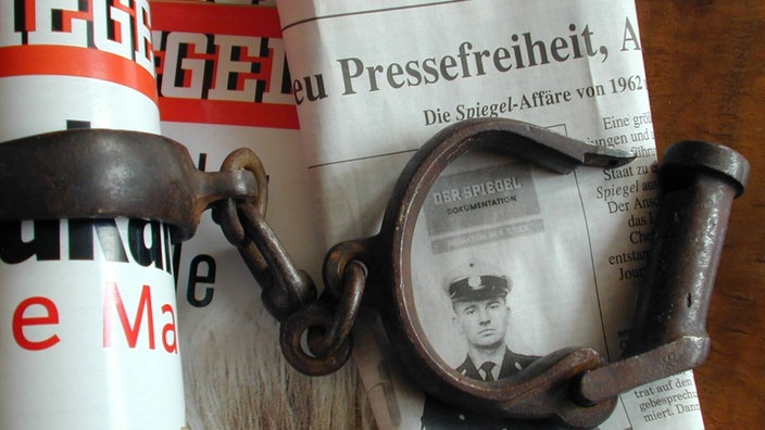 Eine Handschelle liegt auf einer Ausgabe der Zeitschrift "Der Spiegel" und einer Zeitung, auf der das Wort "Pressefreiheit" zu sehen ist