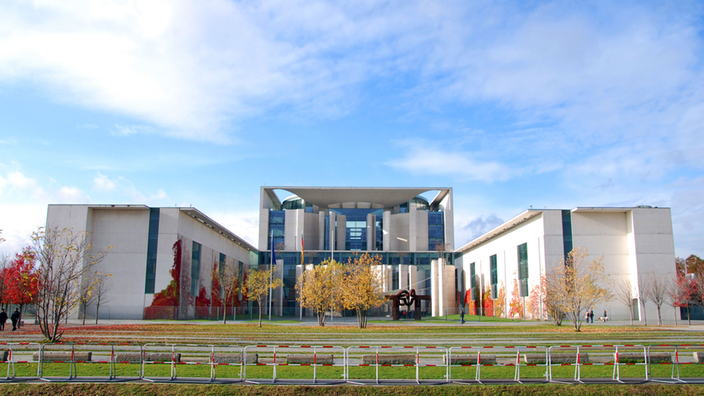 Das Berliner Kanzleramt von weitem: An der Seite vierstöckige Bürospangen, in der Mitte ein achtstöckiger Kubus mit einem halbrunden Riesenfenster; im Hintergrund ein großer Park.