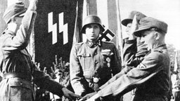 Ein junger Mann in Uniform steht vor einer SS-Fahne und wird vereidigt.