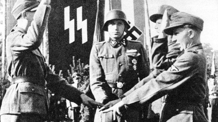 Ein junger Mann in Uniform steht vor einer SS-Fahne und wird vereidigt.