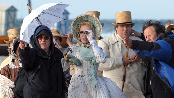 Historisch kostümierte Leute am Strand, in der Mitte eine lockige blonde Dame, die mit dem Opernglas entrüstet ein Geschehen in der Ferne verfolgt.