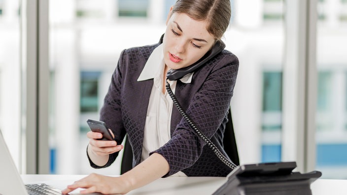 Eine Frau bedient ihren Laptop, ihr Handy und das Telefon gleichzeitig