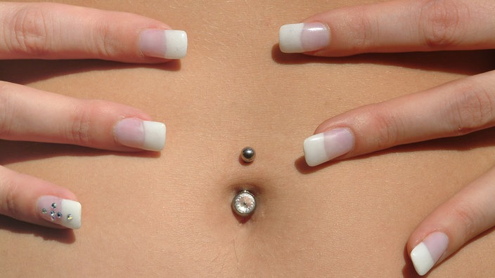 Ein Bauch mit einem Piercing im Bauchnabel, umgeben von jeweils drei Fingern rechts und links.