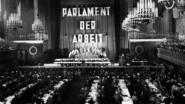 In einer großen Kongresshalle steht an einer Wand in großen Buchstaben "PARLAMENT DER ARBEIT", davor sitzen Gewerkschaftsfunktionäre an langen Tischen