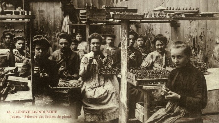 Eine Schwarzweiß-Fotografie im Postkartenformat zeigt einen Raum, in dem von circa zwölf Frauen Zinnsoldaten hergestellt und bemalt werden. Das Foto stammt aus Frankreich und ist auf das Jahr 1900 datiert.