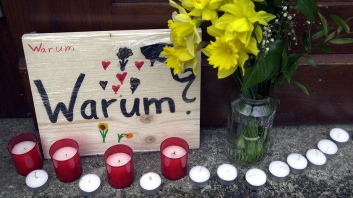 Ein gemaltes Schild mit der Aufschrift "Warum?", davor Kerzen und Blumen.