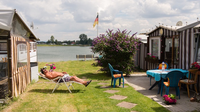 Mann sonnt sich auf einem Campingplatz vor einem bunt gestreiften Zelt auf einem Campingstuhl.