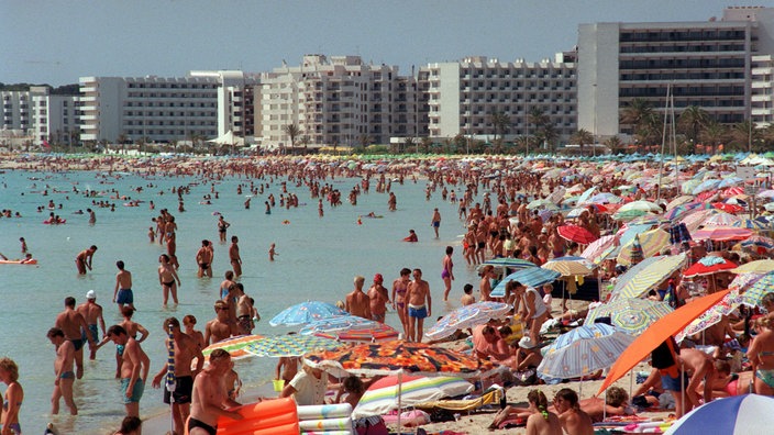 Ein völlig überfüllter Badestrand. Im Hintergrund sind zahlreiche, einheitlich graue Hotels zu sehen.