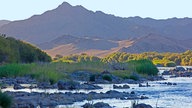 Im Vordergrund ist ein Gewässer mit Steinbänken zu sehen, dahinter ist bergige wüstenähnliche Landschaft.