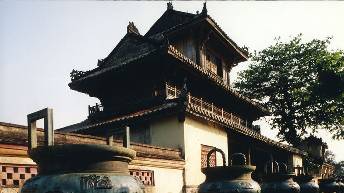 Blick auf ein Gebäude eines vietnamesischen Palastes. Davor ein großer kesselartiger Behälter.