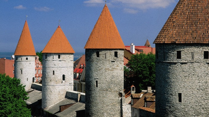 Vier massive graue Türme der Stadtmauer mit spitzen roten Dächern vor blauem Himmel.