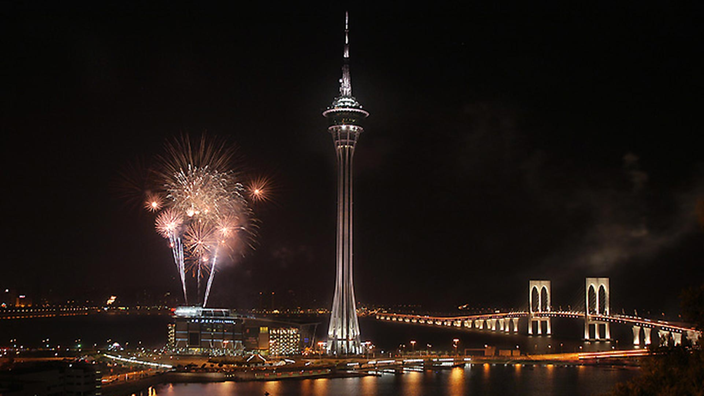 Blick auf die nächtlich beleuchtete Skyline von Macau mit einem großen Turm in der Mitte. Explodierende Feuerwerkskörper sind am Himmel zu sehen.