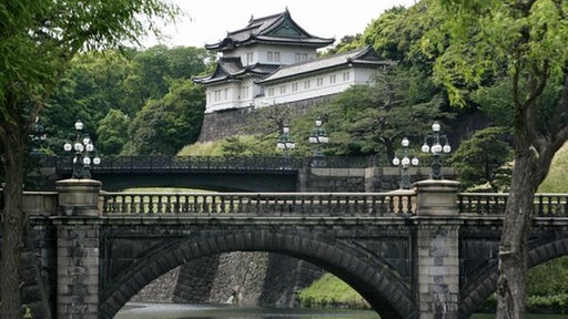 Eine Brücke über einen Fluss, im Hintergrund Bäume und ein Gebäude des Kaiserpalastes mit spitzen, verzierten Dächern.