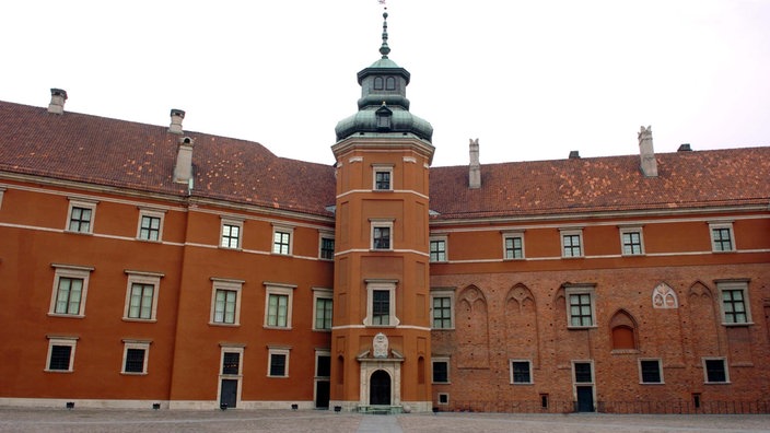 Ein Innenhof wird von beiden Seiten von einem mächtigen Schlossbau aus rotem Stein begrenzt.