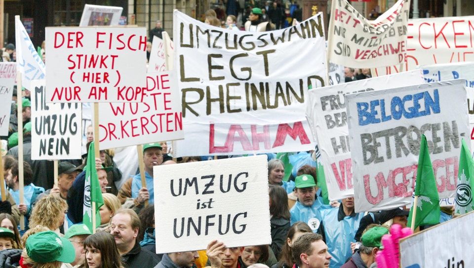 Auf dem Foto sieht man viele Menschen mit Plakaten gegen den Umzug demonstrieren. Auf den Plakaten kann man unter anderem 'Umzug ist Unfug' und 'Umzugswahn legt Rheinland lahm' lesen.