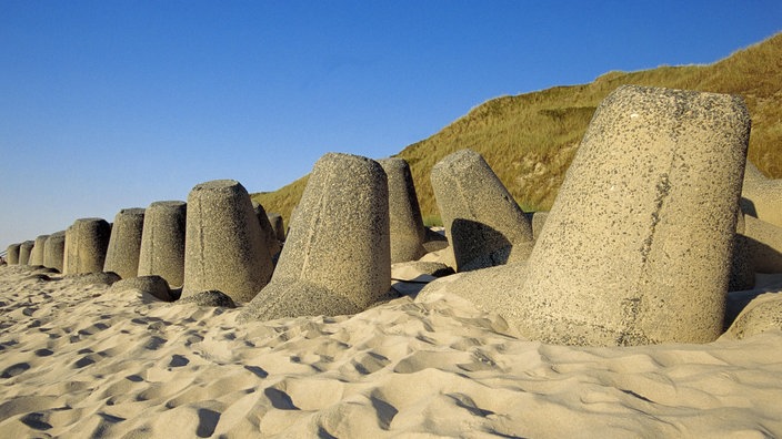 Riesige Betonklötze, die teilweise im Sand versunken sind. Dahinter Dünenlandschaft.