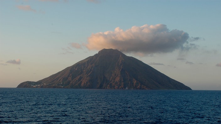 Blick vom Meer aus auf die Vulkaninsel Stromboli im Abendlicht. Rauchschwaden steigen über dem Krater auf.