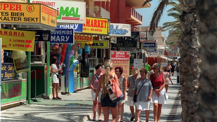 Einkaufsstraße in El Arenal. Rechts gehen Passanten, links sieht man die Schilder der Läden wie 'Kalter Sekt', 'Sangria' oder 'Souvenirs'.