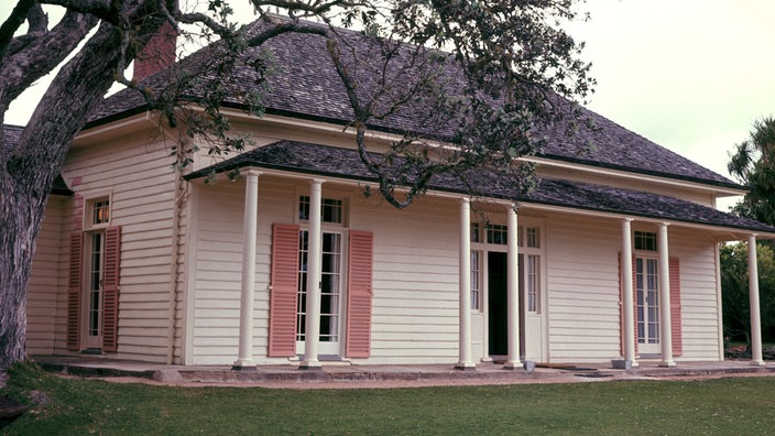 Das Bild zeigt ein relativ kleines Haus aus weißem Holz mit schwarzem Dach. Es hat ein kleines Vordach, das ebenfalls schwarz ist. Die Fensterläden sind lachsfarben. Links neben dem Haus steht ein großer Baum.