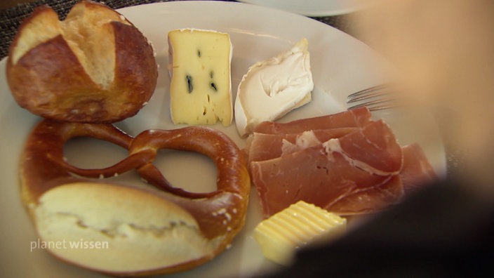 Teller auf dem ein Laugenbrötchen, eine Brezel, sowie Schinken, Käse und Butter liegt.