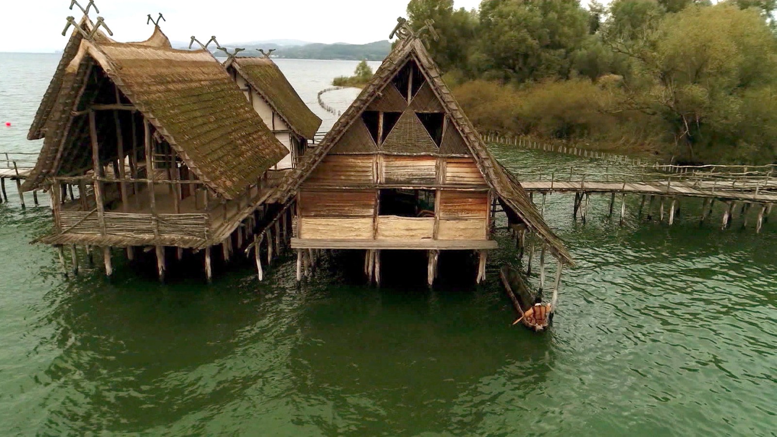 Steinzeit-Pfahlbauhäuser stehen in einem See per Steg mit dem Land verbunden.