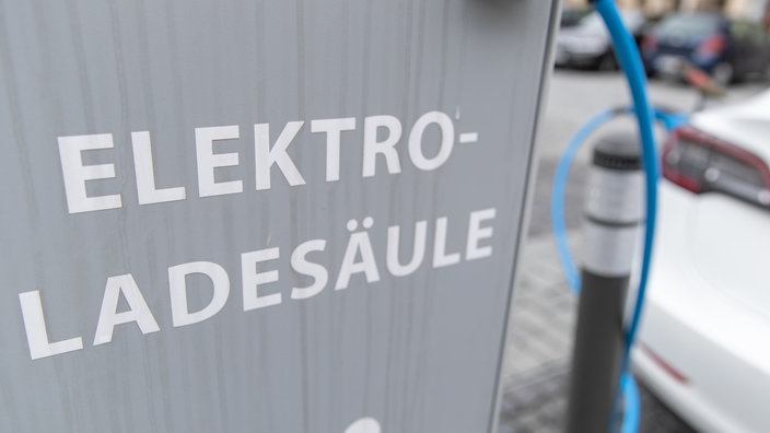 Graue Elektro-Ladesäule. Im Hintergrund weißes Elektroauto