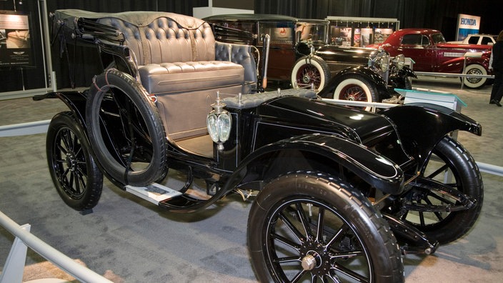 Ein schwarzer Oldtimer steht in einer Museumshalle. Er hat beige Ledersitze und ist offen, wie eine Kutsche